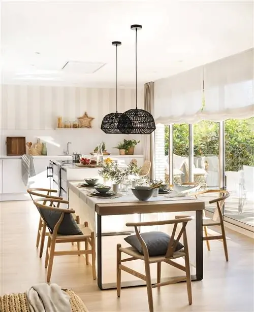 Открытая белая кухня от Pia Capdevila с островом и кабинетом с большим деревянным столом и потолочными светильниками.