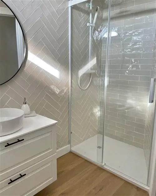 Ванная комната после ремонта с душем с экраном, деревянным полом, белым шкафом и круглым зеркалом.