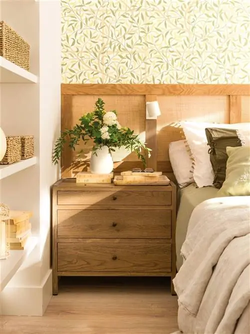 Спальня с деревянным изголовьем и сеткой на обоях с ботаническими мотивами и деревянной тумбочкой.