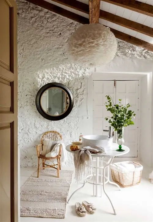 Необработанная каменная стена, выкрашенная в белый цвет, украшенная круглым зеркалом в черной раме.