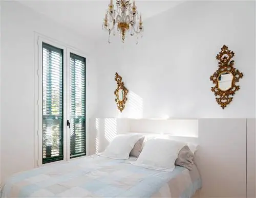 Главная спальня после ремонта с белым изголовьем, окном, золотыми зеркалами и люстрой.