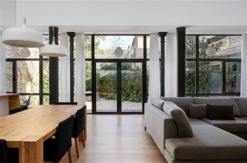 Гостиная после ремонта с черными окнами, стеклянной дверью, деревянным обеденным столом с черными стульями и серым диваном.