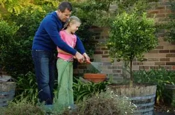 Отец и дочь (10-12 лет) поливают сад из шланга, вид сбоку