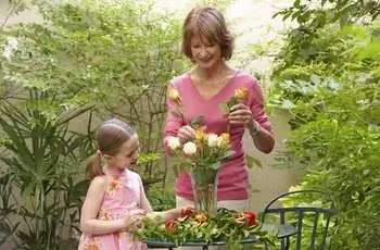 Бабушка и внучка (6-8 лет) расставляют цветы в вазе и улыбаются.