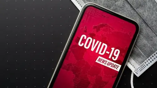 Телефон в серой хирургической маске отображает красный экран с белым текстом об обновлении новостей о COVID-19.