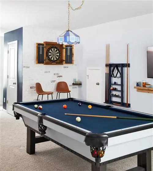 бильярдный стол из синего войлока со светло-голубой люстрой вокруг него и доской для дартс на задней стене из белого кирпича