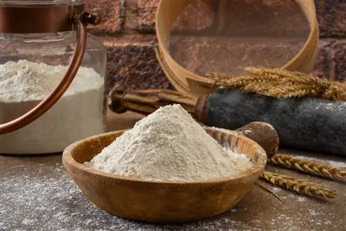 Миска муки на столешнице со скалкой и стеклянной тарой на заднем плане