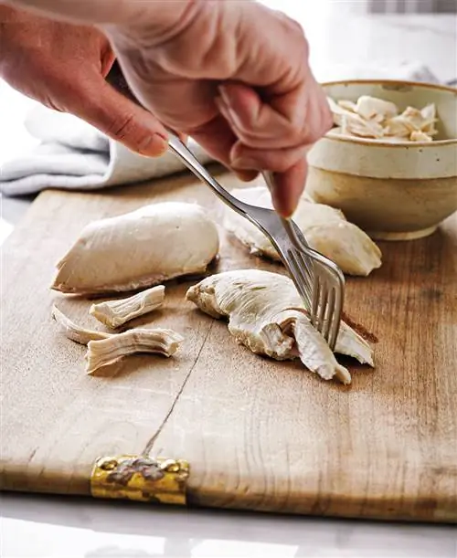 Две руки измельчают курицу вилками на деревянной разделочной доске с миской на заднем плане