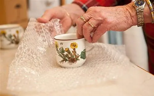 Пожилая женщина упаковывает прекрасную фарфоровую чашку в пузырчатую пленку, прежде чем переехать в свой новый дом.