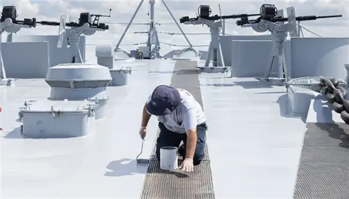 На этом изображении изображен человек, стоящий на четвереньках, с малярным валиком, красящий нос корабля.