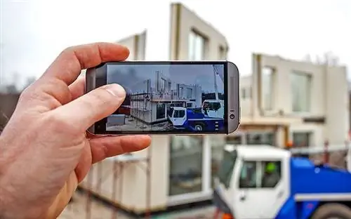 Домовладелец держит телефон с камерой, фотографируя свой новый модульный дом, установленный на заднем плане. Домовладелец держит телефон с камерой, фотографируя свой новый модульный дом, установленный на заднем плане.