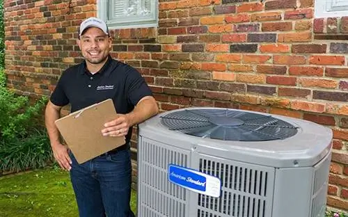 Дружелюбный техник американского стандарта HVAC, который носит кепку, улыбается, держит планшет и стоит рядом с конденсаторным блоком.